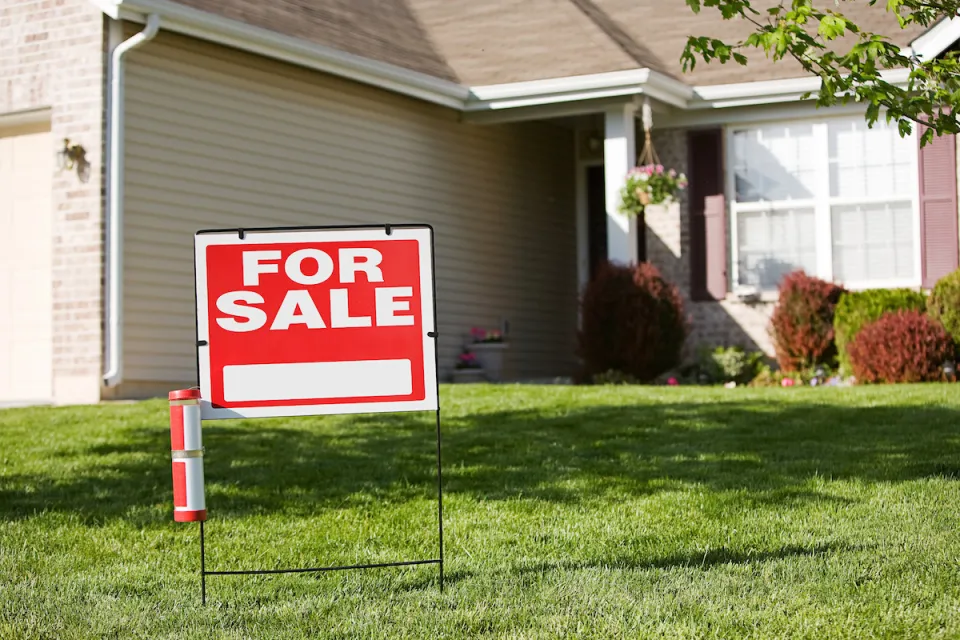 Red and white "For Sale" sign in front yard of suburban house.